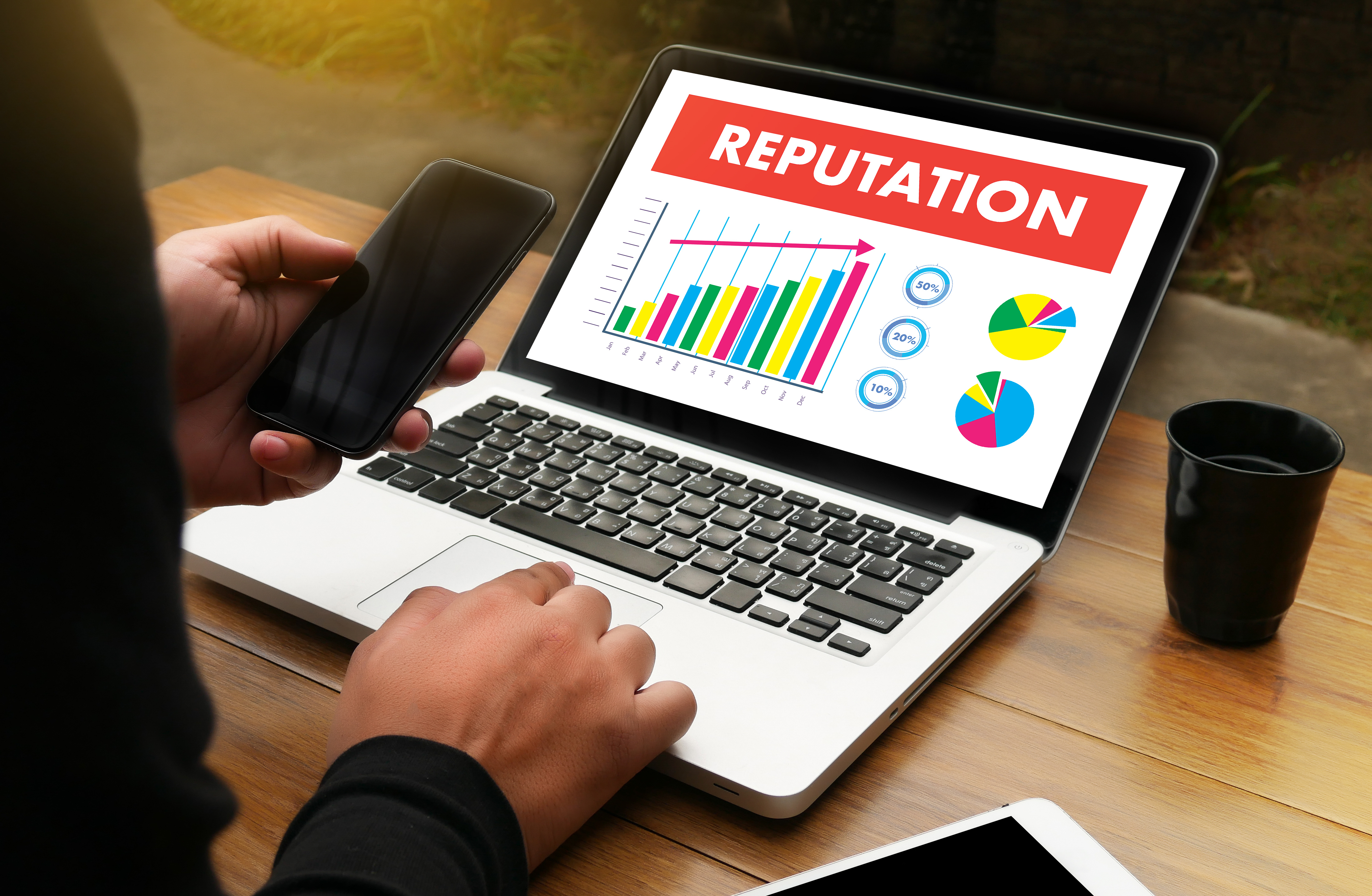 a close up of a mans arms sitting at a desk with a laptop, he is wearing a suit and working on the laptop, he has his cellphone, face-up, in his left hand. The computer screen has a graph with a trend line incrementally growing. The graph is titled Reputation