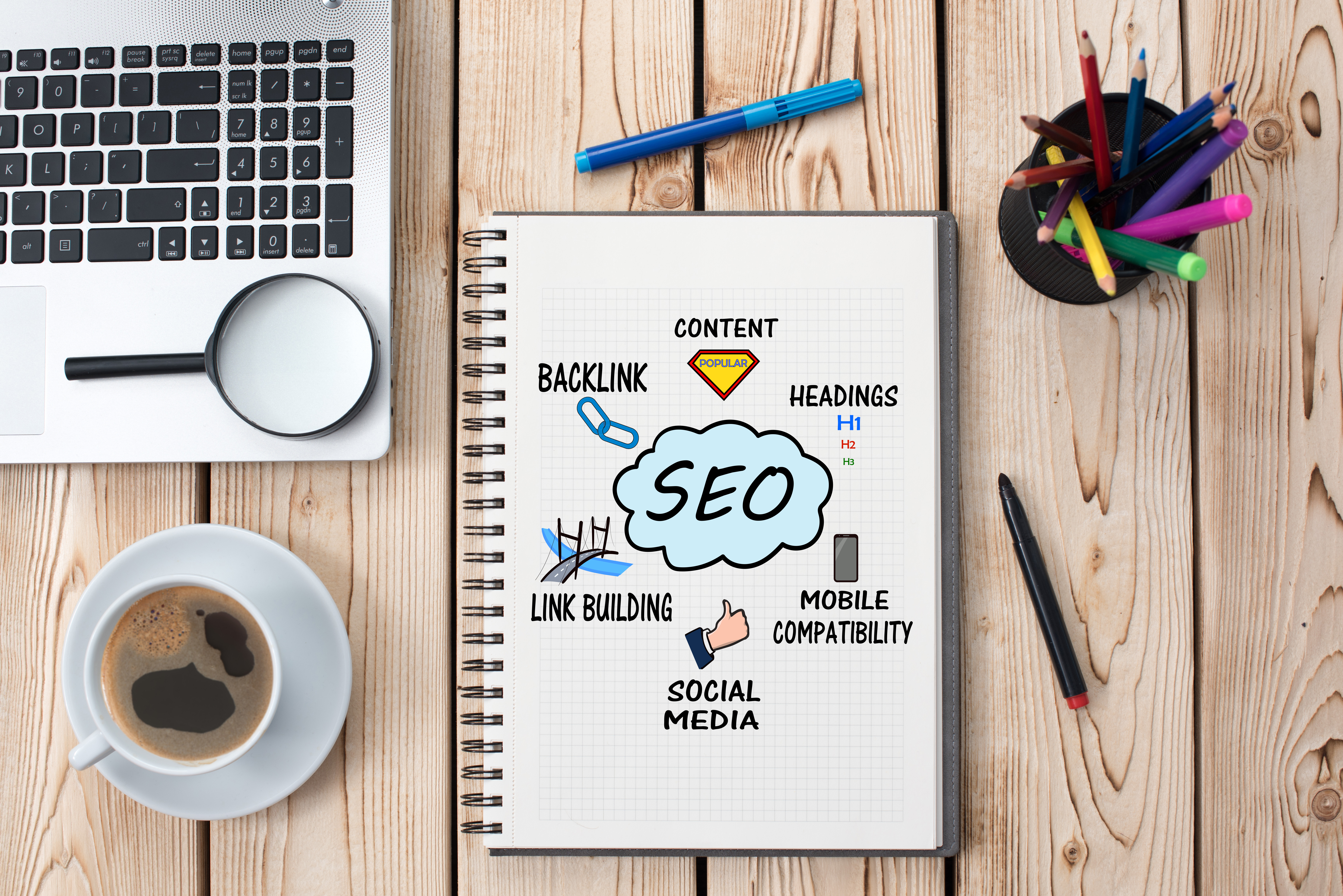 birds eye view of a table, a laptop with a magnifying glass, a cup of black coffee and a cup of pens and pencils surround a notebook. In the notebook SEO is written in a cloud, around it content, backlink, headings, link build, mobile compatibility and social media is written 
            next to images that describe these titles