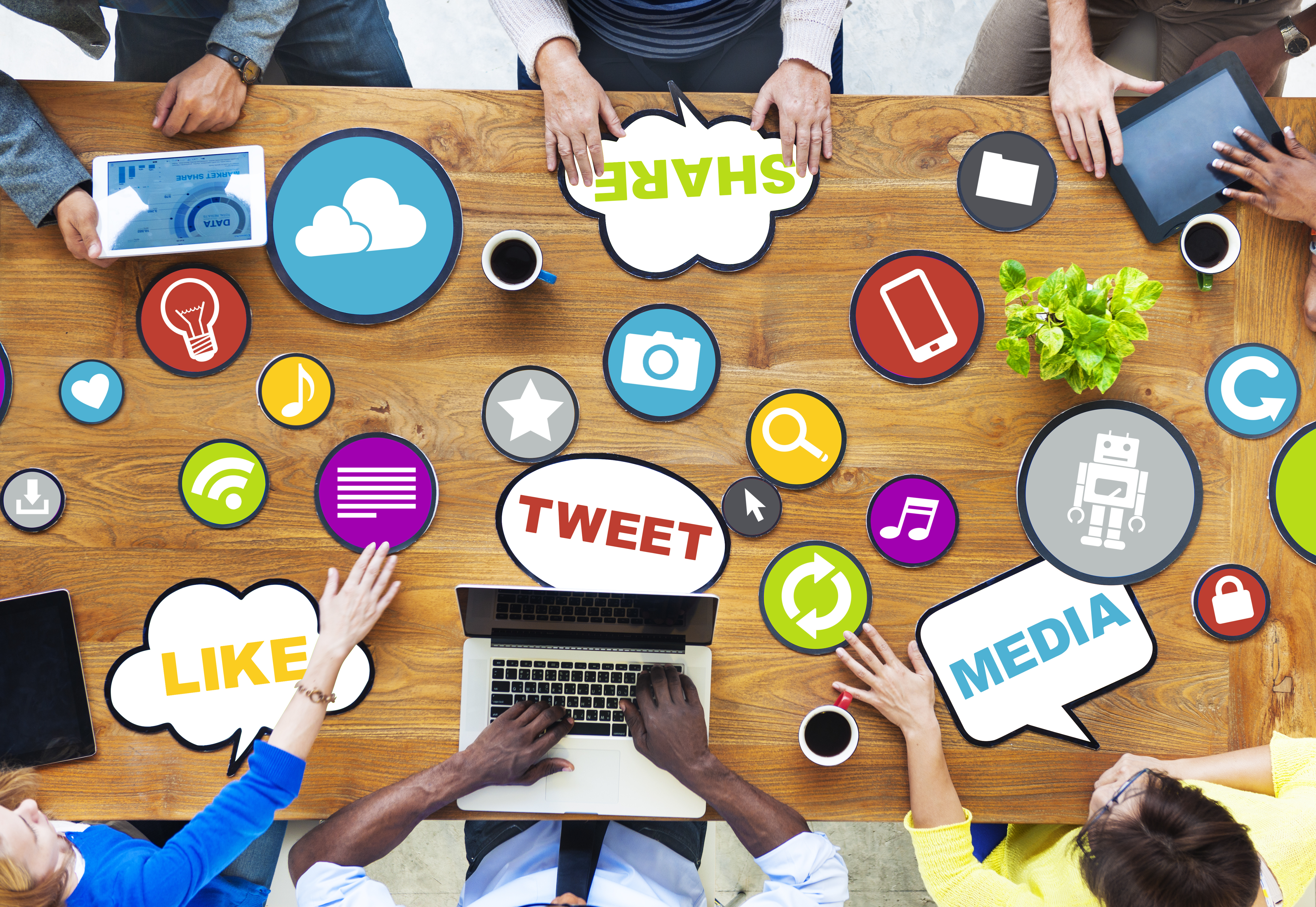 Birds eye view of a large conference table with 6 workers sitting around it. There are many brightly coloured placards with symbols on them scatered aroudn the table, some tought bubbles, some circles. There are also multiple devides such as ipads and laptops as well as cups of coffee